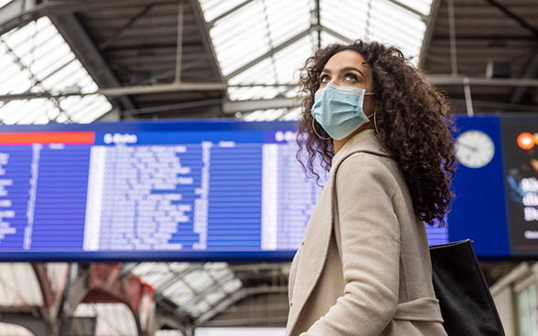 Eine Reisende mit Maske vor einem elektronischen Fahrplantafel in einem Bahnhof